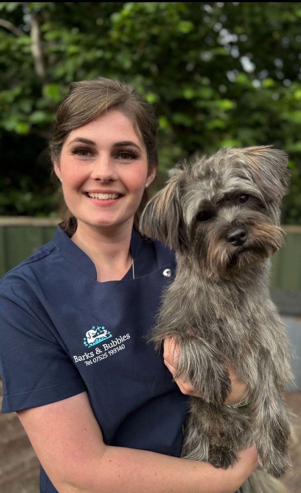 The Barks & Bubbles groomer, in a navy Barks & Bubbles tunic, holding a grey shaggy terrier
